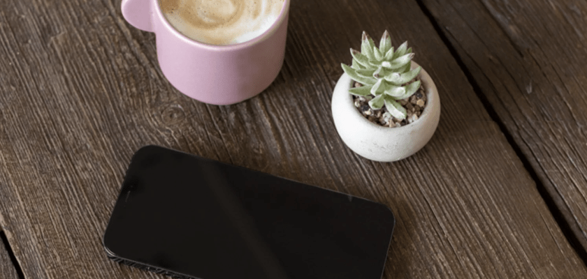 iPhone mockup on a wooden table with a coffee cup and plant, ready for your design