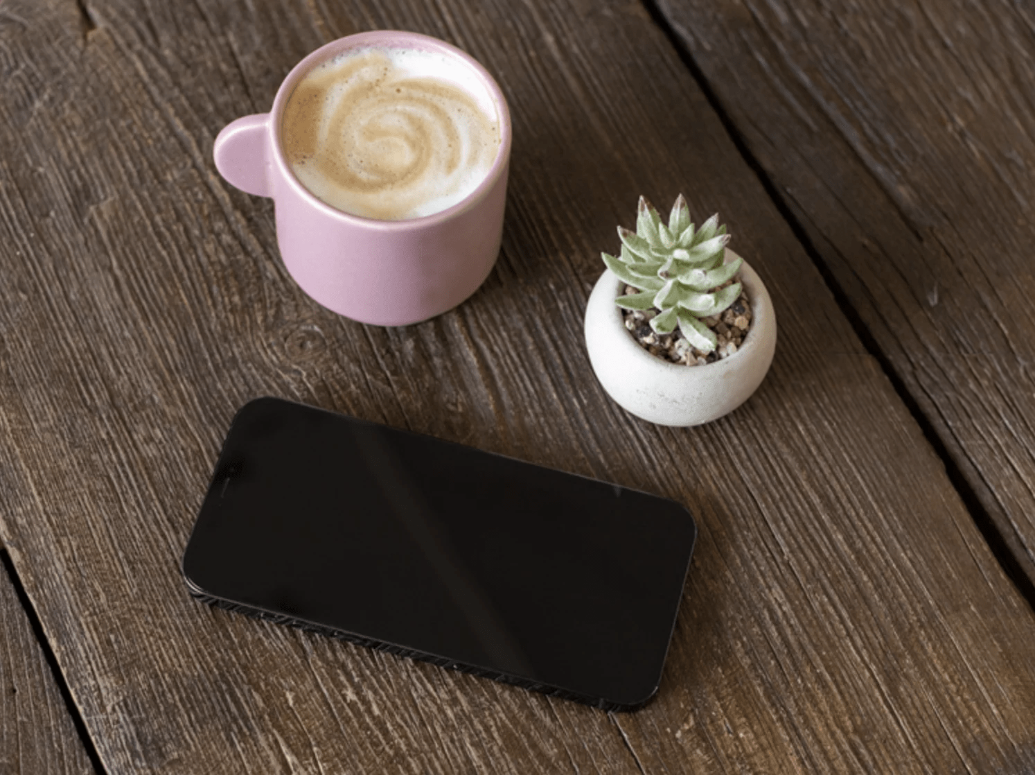 iPhone mockup on a wooden table with a coffee cup and plant, ready for your design