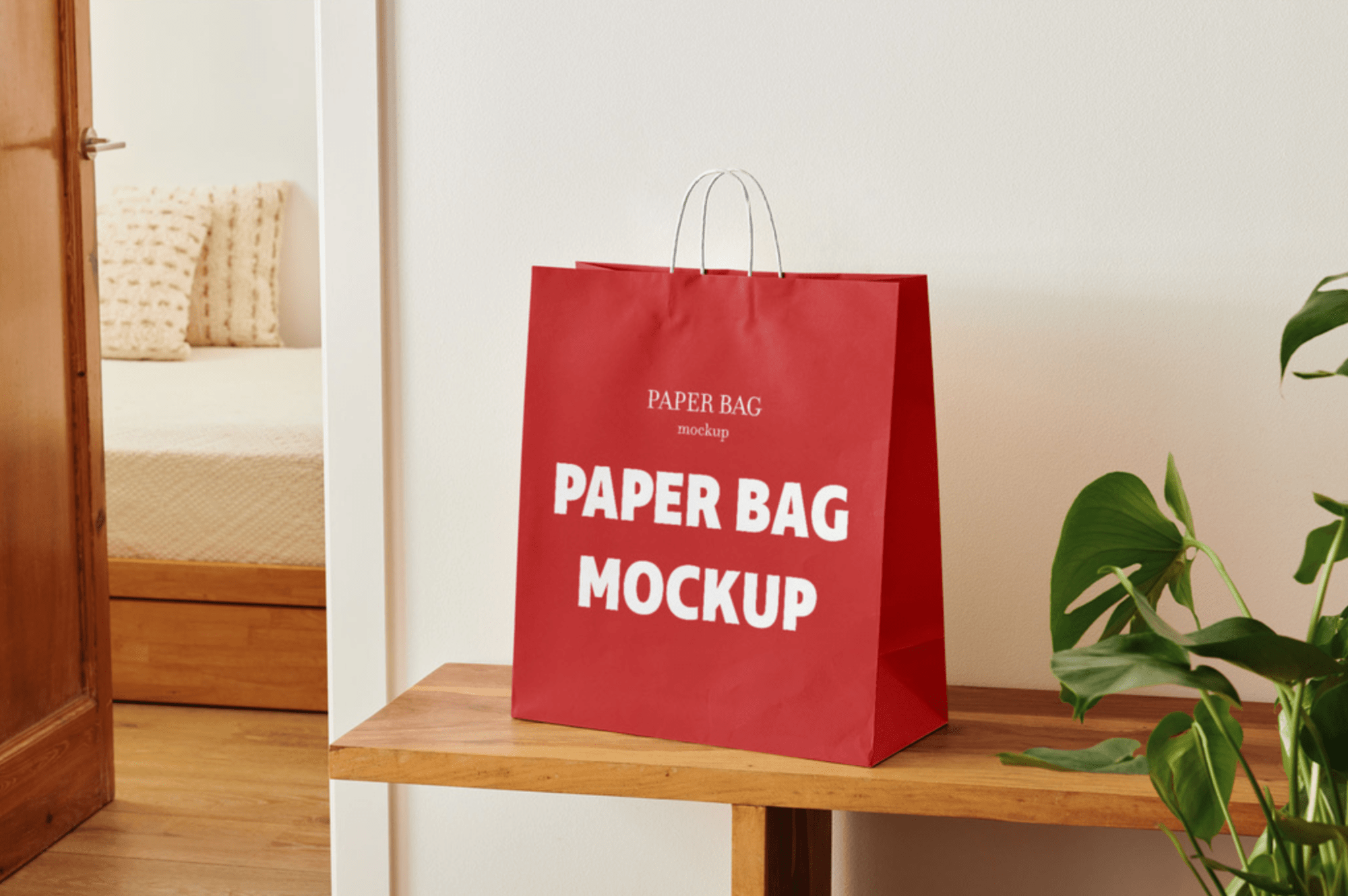Red paper shopping bag mockup placed on a wooden bench with a white wall background and a green plant to the side.