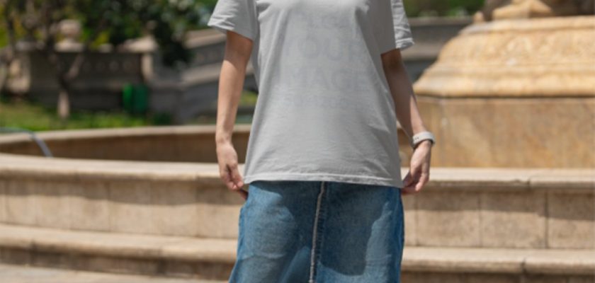 Free apparel mockup of a woman in a Bella Canvas t-shirt and denim skirt standing by a fountain