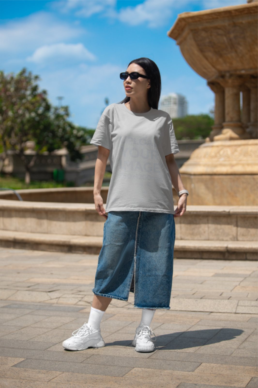 Free apparel mockup of a woman in a Bella Canvas t-shirt and denim skirt standing by a fountain